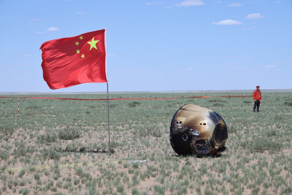 The retrieval site of the capsule of the Chang'e-6 probe in Siziwang Banner, north China's Inner Mongolia. China's flag is on the left of the probe capsule which appears on the ground on its side, with heat marks on the exterior. A man in red stand near a cordon tape in the background
