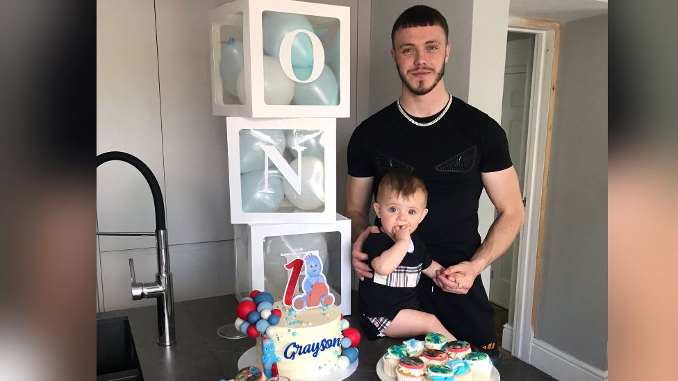 Reece Thomas Thompson pictured with his son Grayson on the boy's first birthday, with a cake next to them and cubes full of balloons spelling ONE