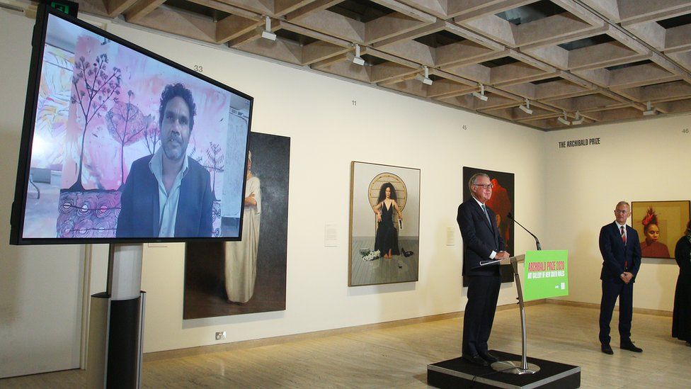 Vincent Namatjira seen on screen behind gallery director Michael Brand at the announcement of the prize