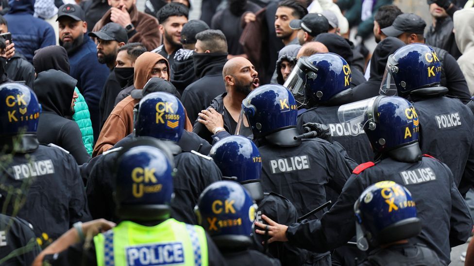 A group of angry-looking men dressed in mainly black clothes and wearing face coverings stand in front of a group of police wearing riot gear and holding batons