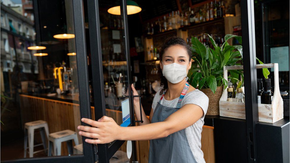 A happy business owner opening the door at a cafe while wearing a facemask