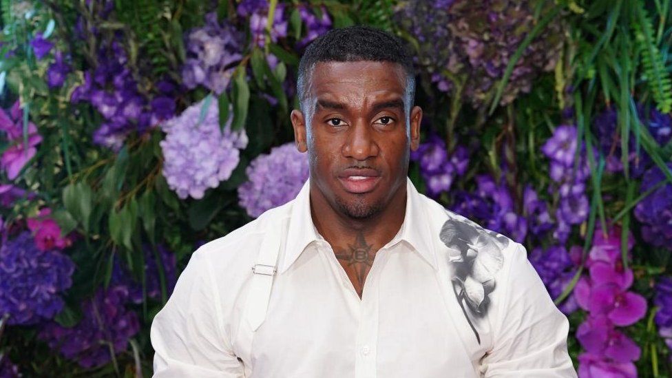 Bugzy Malone on the red carpet gala of #TheMikeGala which was hosted by Stormzy at The Biltmore Mayfair in London. He is looking straight at the camera and isn't smiling. he has short dark hair and a bit of stubble around his chin and on his top lip. He is wearing a white shirt that is buttoned up to near the top and there is a black floral pattern on his left shoulder. Behind him is a wall of purple flowers and green leaves.