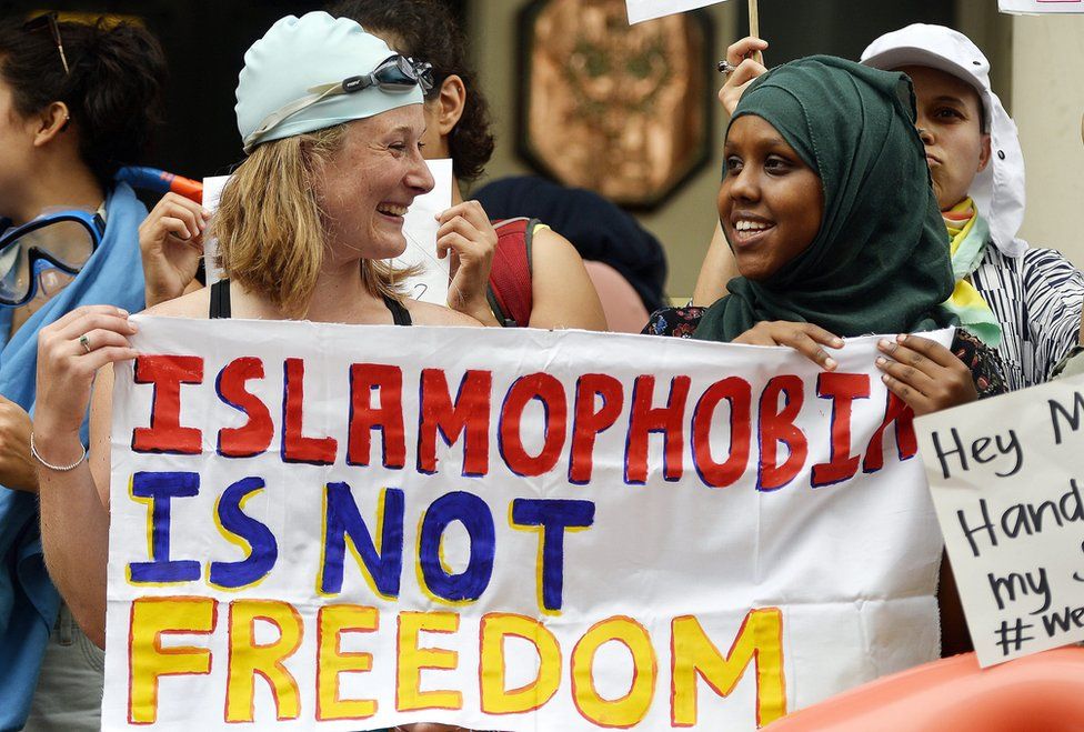 Two women - one white and in a swimming cap and goggles, the other black and wearing hijab - hold a sign saying 'Islamophobia is not freedom'
