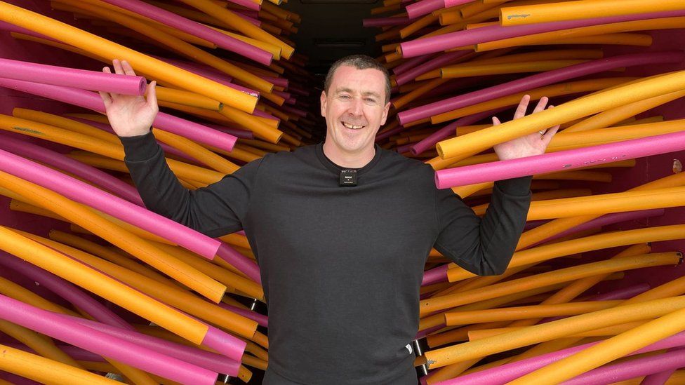A man in a black long-sleeved t-shirt stands between two dense rows of pink and yellow tubing. They run from the top to the bottom of the frame and extend far back out of site. He's smiling and has his hands up, palms facing forward, as if resting on the tubes.