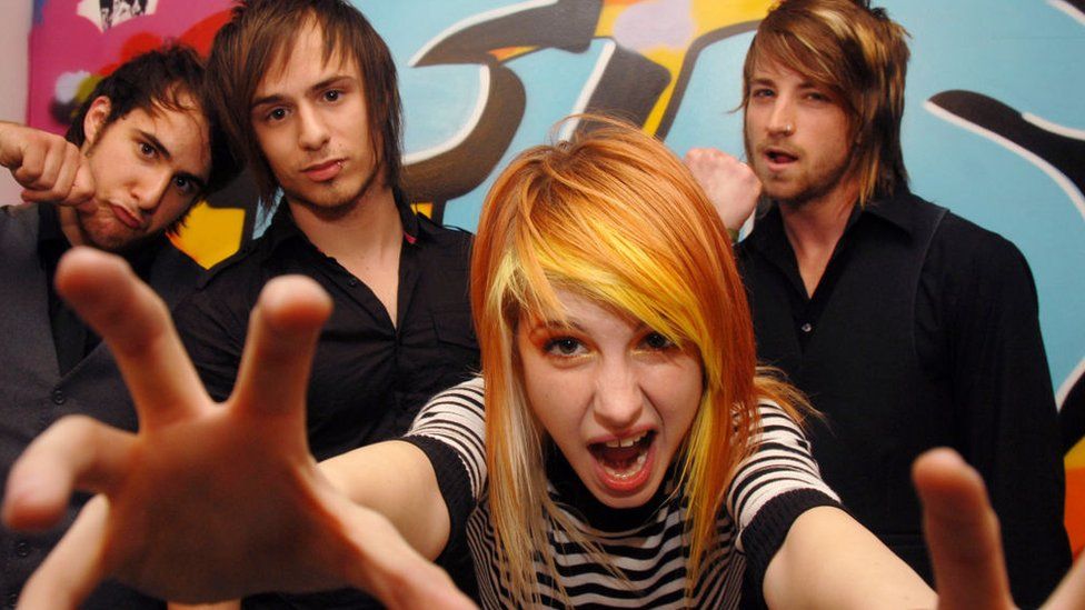 Paramore pictured in 2007. Singer Hayley Williams has dyed orange hair and wears a striped black and white top, her arms outstretched towards the camera. Her bandmates have long fringed hair and are pictured in front of a graffitied wall.