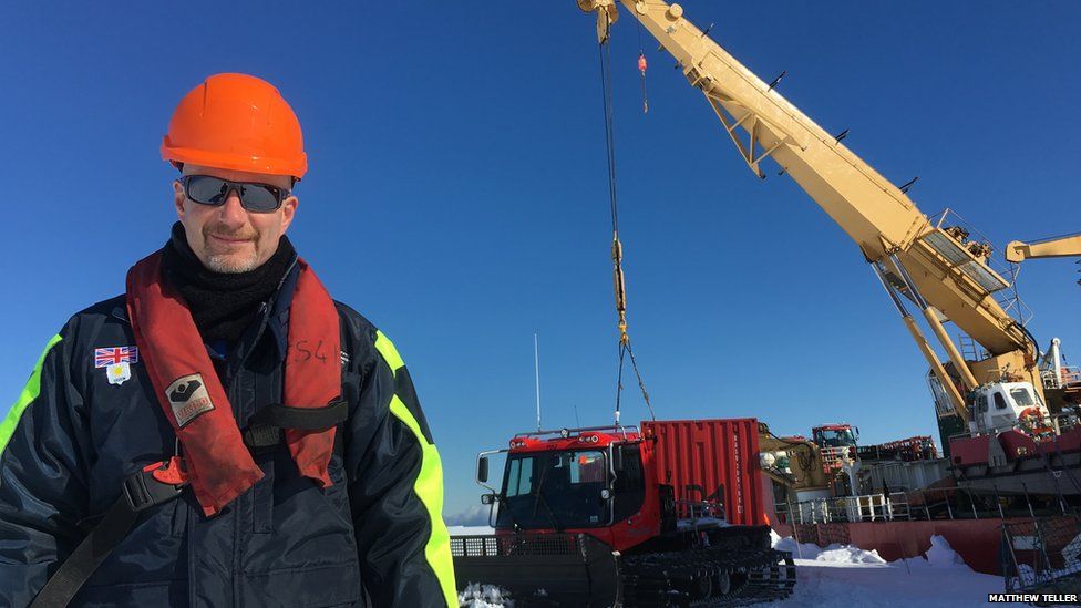 Peter Gibbs in foreground dressed in warm winter clothes. Standing on white snow and bright blue sky behind. Large ship and crane in background.