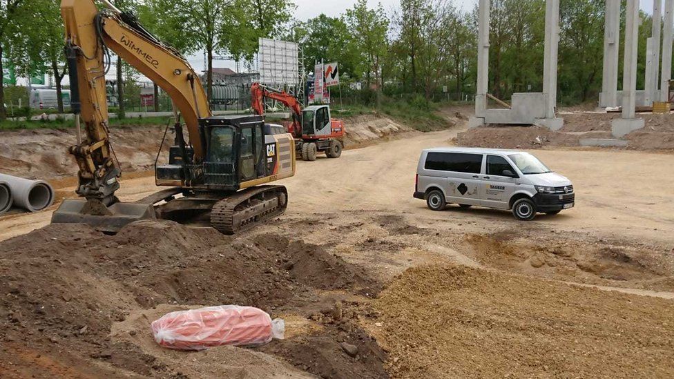 Виден строительный экскаватор, неподвижный и пустой, с копающей рукой рядом с неразорвавшейся бомбой, завернутой в оранжевый пластик