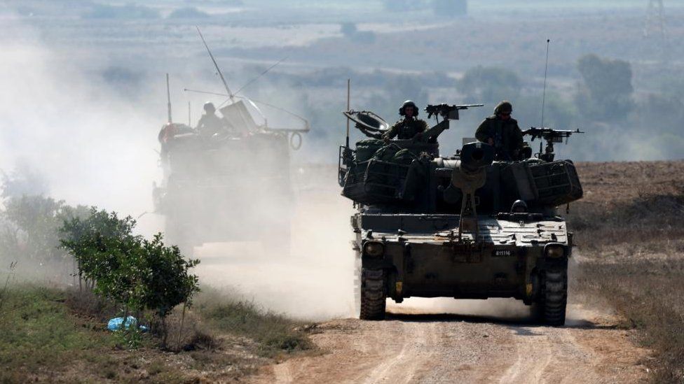 Two Israeli tanks seen moving along a duty road with soldiers perched on top of them.