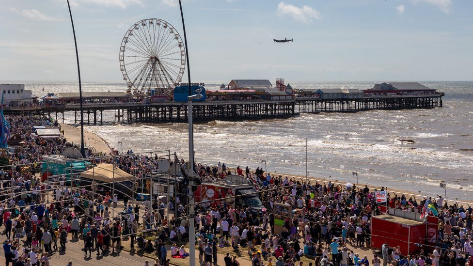 Crowds of spectators throng along Blackpool Promenade and the pier as a Lancaster Bomber flies overhead