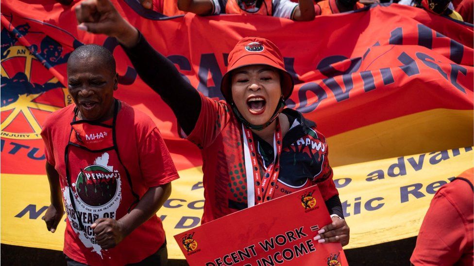 A woman at a protest in Johannesburg standing next to man and a crowd in the background. The people are wearing all red. The woman has her fist in the air and appears to be shouting. She is carrying a banner which says: "decent work equals fair income."