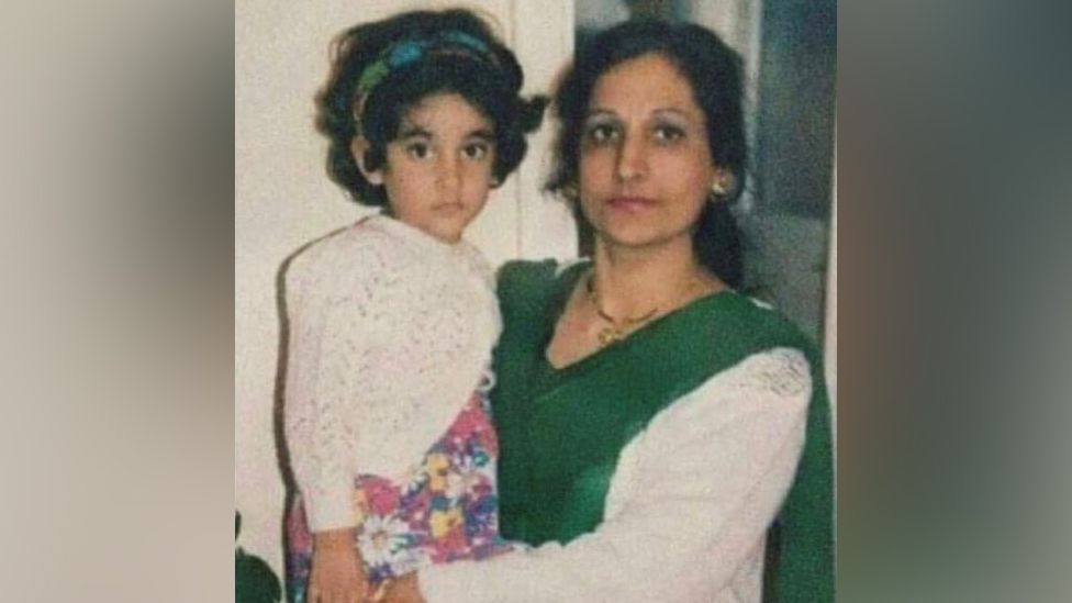 An older picture of Sonia and her mum. Sonia is a child, and is being lifted by her mum in her hands. Sonia is wearing a white knitted sweater and flowery dress. Her mum is wearing a white top with green shawl.