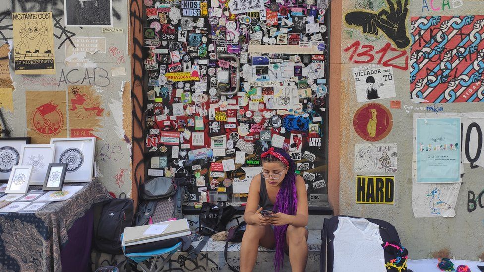 A woman sits in front of a wall covered in stickers and graffiti in Santiago