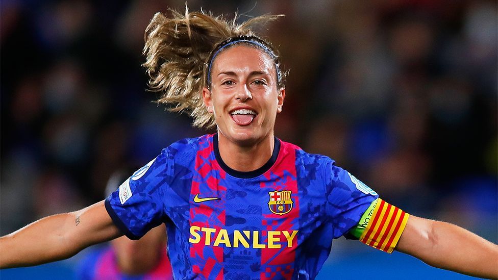 Alexia Putellas, a female footballer in a blue Barcelona jersey with a red stripe pattern. Her arms are outstretched in celebration.