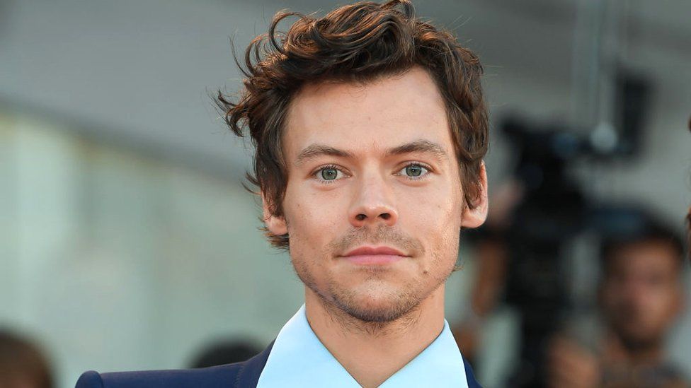 Harry Styles before the haircut. Harry is a 29-year-old white man with curly brown hair. He has a light stubble and blue eyes. He is pictured outside at an event wearing a pale blue shirt and navy blazer