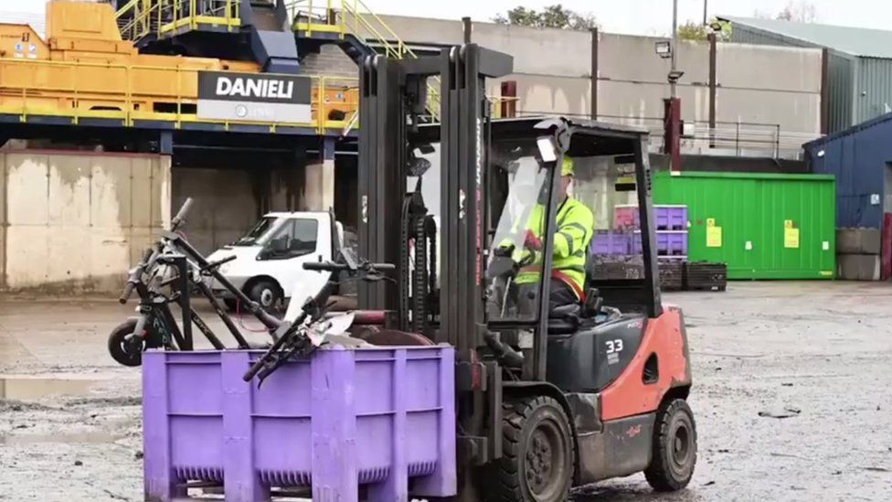 Man in high viz drives a fork lift truck which is carrying a purple crate containing e-scooters