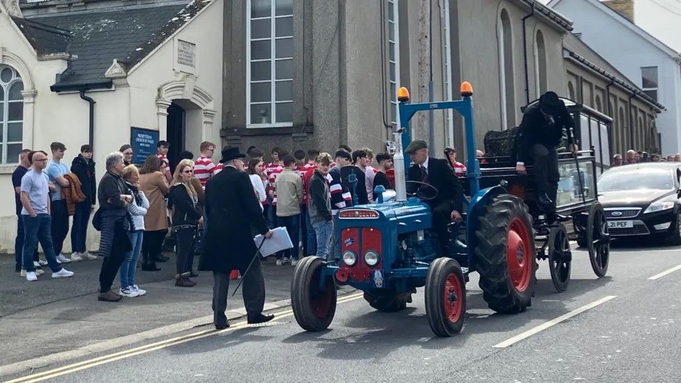 a blue tractor carries the 16-year-old boy's coffin through the village