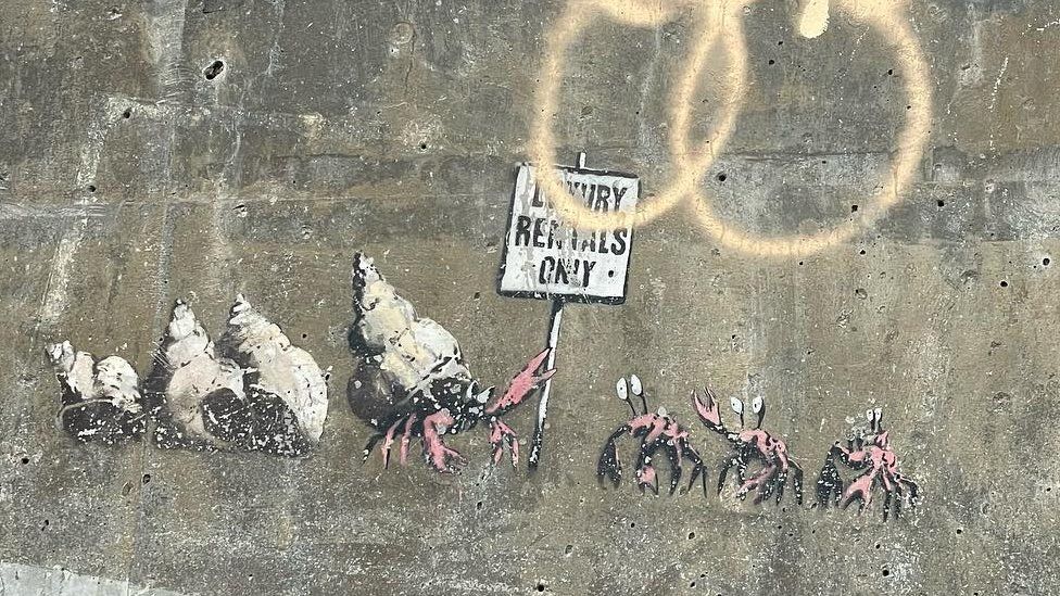 Banksy mural of hermit crabs, with spraypaint covering some of the sign held by one of the hermit crabs, which reads "Luxury Rentals Only"