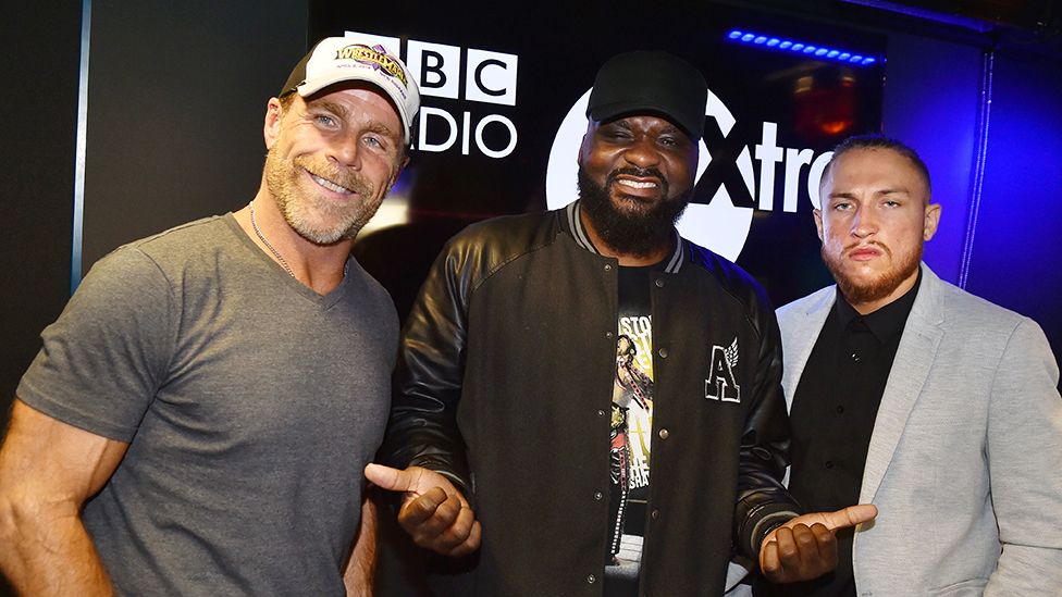 DJ Ace with WWE legend Shawn Michaels (left) who is wearing a grey tshirt, silver chain and cap and British wrestling star Pete Dunne (right) who is wearing a black top under a light grey blazer. Behind them is a black screen with "BBC 1Xtra" written in white.