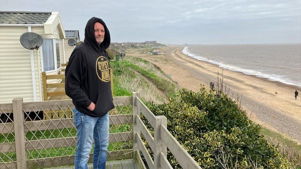 Man with grey beard wearing dark-coloured hoodie standing in a garden on the cliff edge