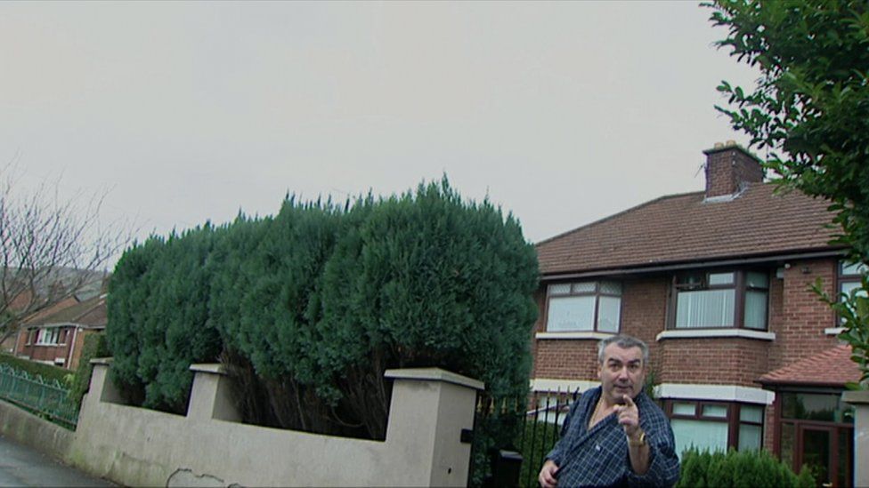 Freddie Scappaticci pointing at the camera with his eyebrows raised. Semi-detached houses and a hedge are visible behind him.