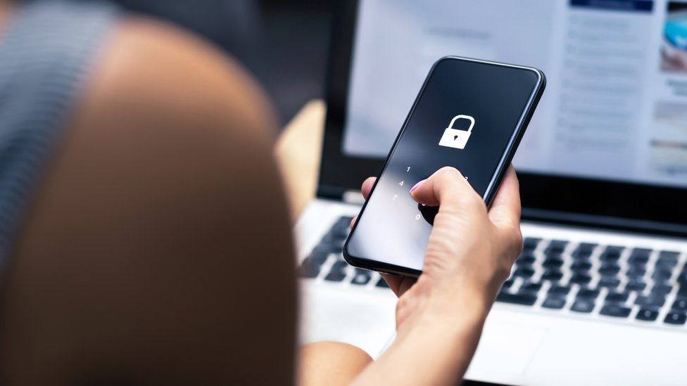 Stock image of a woman unlocking a smartphone while seated in front of her laptop