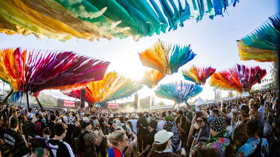 A crowd of festival goers gathers underneath several rainbow-coloured installations that resemble upturned umbrellas. The mass of people stretches off into the distance, with large screens and a festival stage visible on the horizon. The scene is illuminated by blazing sunshine.