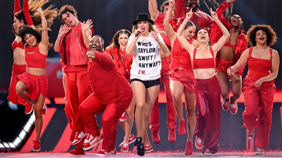 Taylor Swift in a "Who's Taylor Swift anyway?" t-shirt with dancers dressed in red outfits