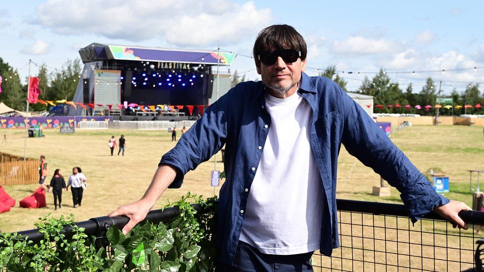 Alex James poses in front of a stage at the Big Feastival, wearing sunglasses