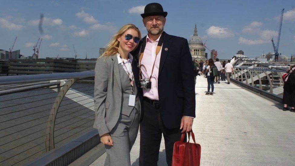 A man and woman stand on millennium bridge. Man is in a top hat and suit, woman is in a suit.