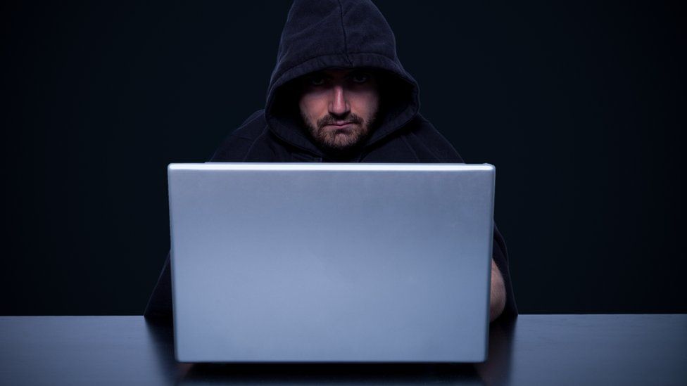 Image of a man in a dark hood sitting at a computer.