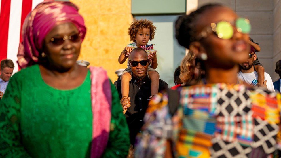 A man wearing sunglasses watches on with a young boy on his shoulders. There are two women in front of him both wearing sunglasses. One is wearing traditional clothing - Dakar, Senegal, Saturday 20 November 2021