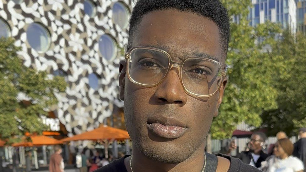 Toye, an Asake fan pictured outside the O2 Arena in London ahead of the singer's first UK show since the Brixton crush. Toye, a black man in his 20s, is looking at the camera with a serious expression. He wears wide round glasses with a clear brown frame and has his hair trimmed short. He wears a black T-shirt and a bag across his body. He is pictured outside in front of trees and a building with round windows