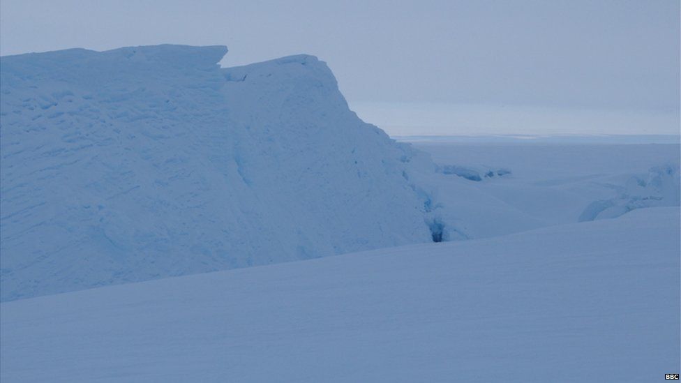 A landscape of pure white snow, with a few dips in the surface and a big mound on the left hand side. The sky is white.
