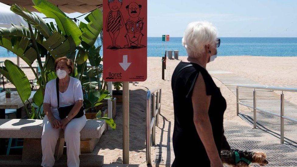 A woman walks her dog past another woman sitting on a Spanish beach