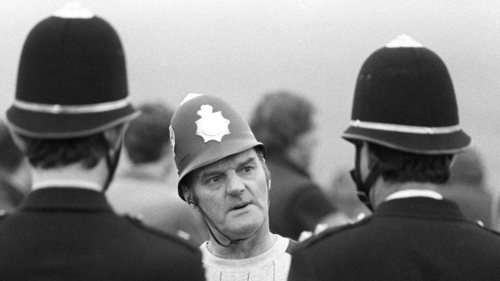 A man wearing a toy police helmet looks at two policemen