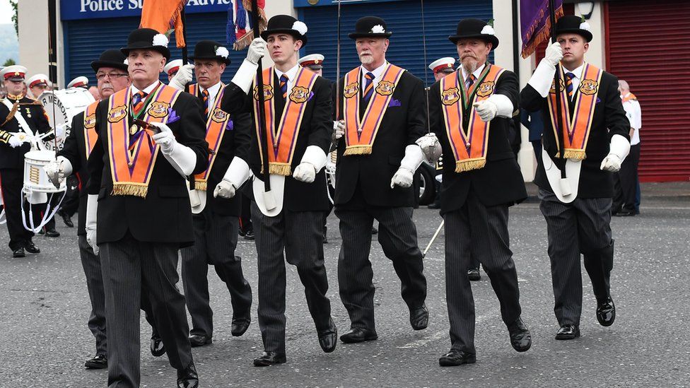 A group of Orangemen are marching wearing orange sashes, white gloves and bowler hats