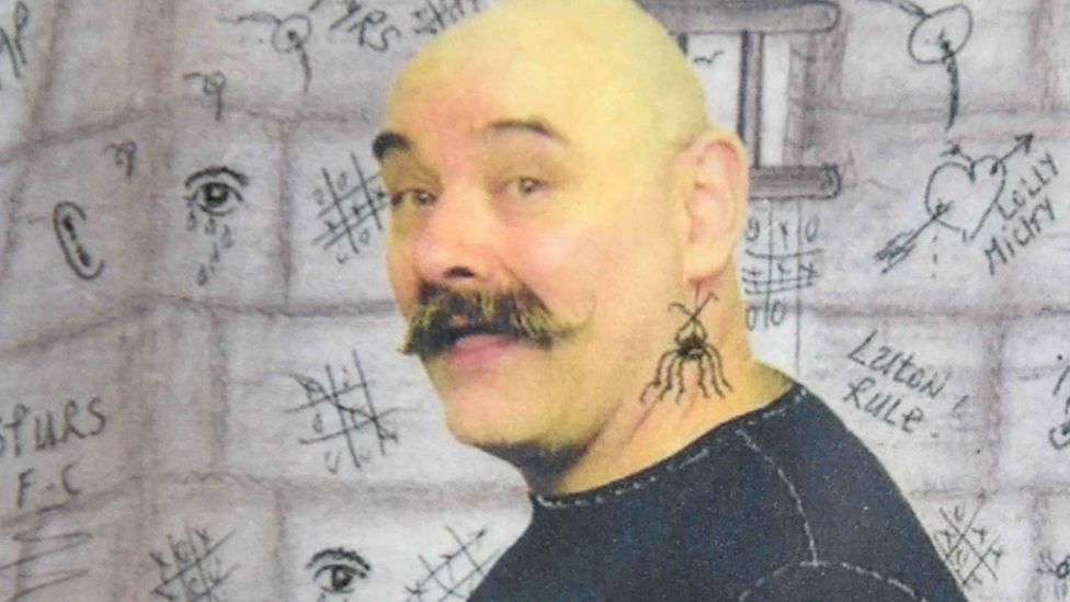 A photo of Charles Bronson turning to the face the camera. He is wearing a black top and has a spider tattoo below his left ear. The background image appears to be a sketch of doodles on a cell wall, including noughts and crosses and a heart with an arrow through it