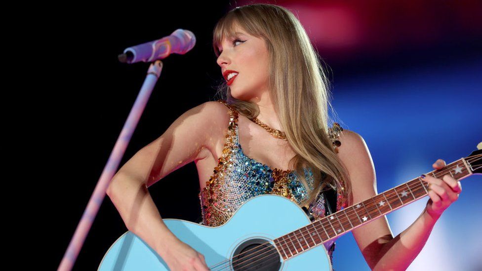Taylor Swift on stage during her Eras tour. She's holding an acoustic guitar with a pale blue body and fretboard with matching stars at key points down to the sound hold. She's in front of a white microphone stand which has taken on a multicoloured hue from the various stage lights. She's wearing a gold, rope-style necklace and a sequined dress that's blue and gold in colour. She's looking to the side, giving a slight smile. Her hair is straight and she's wearing red lipstick.