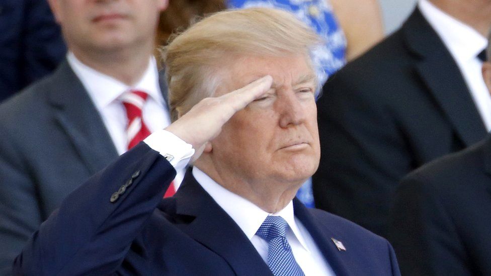 Donald Trump salutes, standing, from a viewing platform with other dignatories
