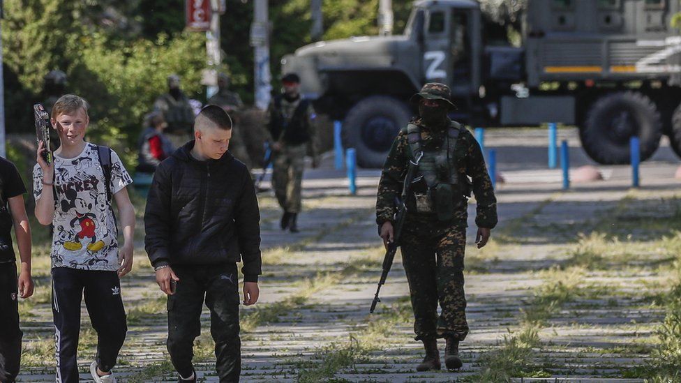 Two boys walking on a road with armed soldiers behind them, and an armoured vehicle marked with 'Z' in the background