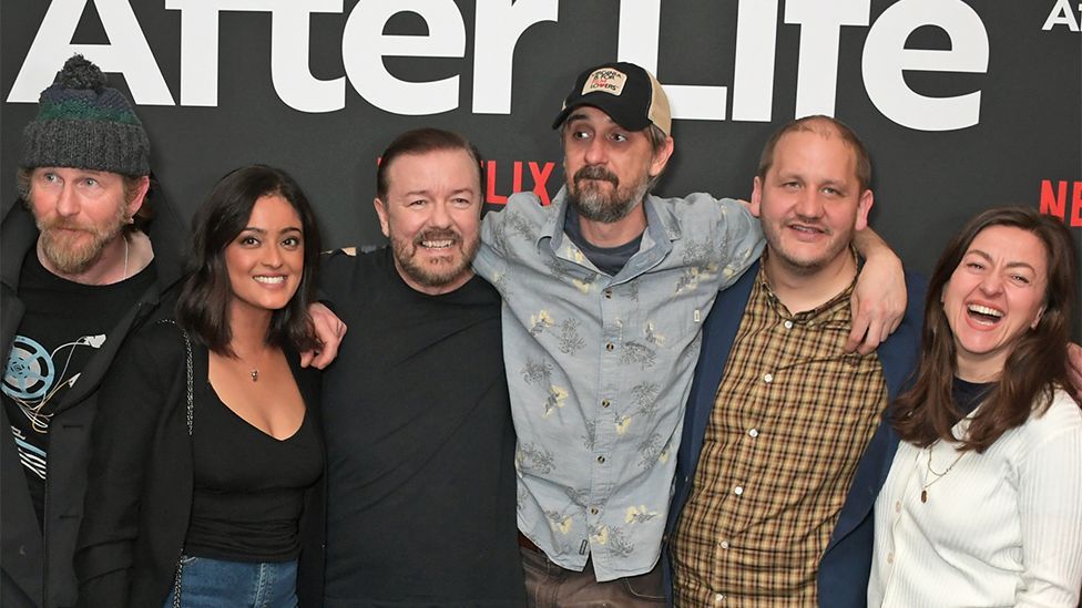 The cast of After Life at a launch event. Left to right is a man, woman, man, man, man and woman. The background has Netflix branding.