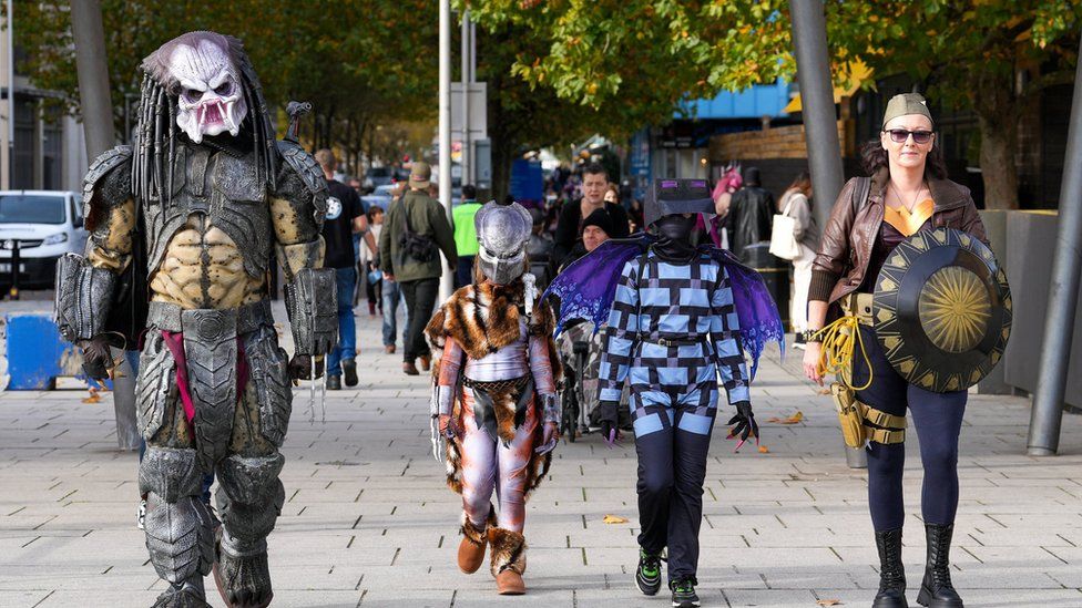A man dressed as the Predator and woman dressed as Wonder Woman walk with two children in costume