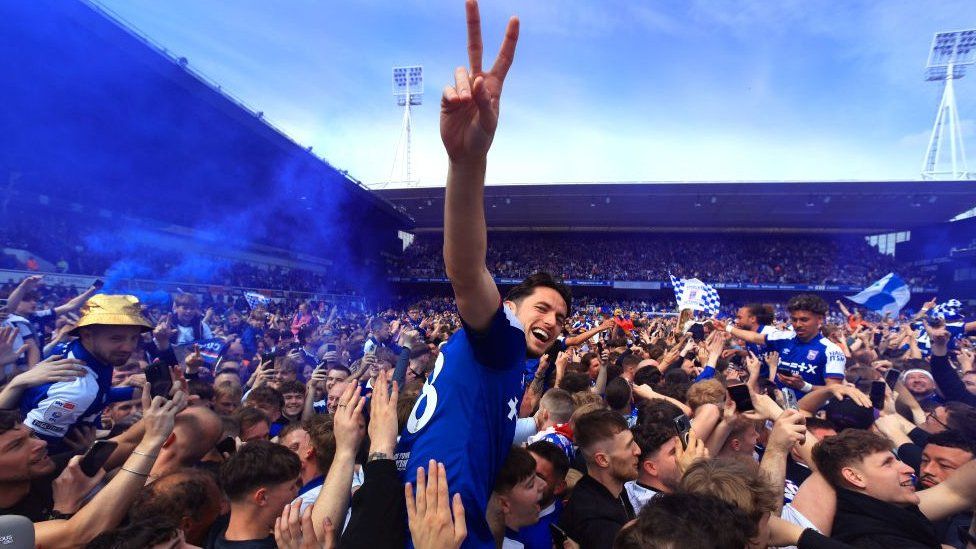 Lewis Travis, player for Ipswich Town, on the shoulders of fans in the crown of the stadium making a peace sign