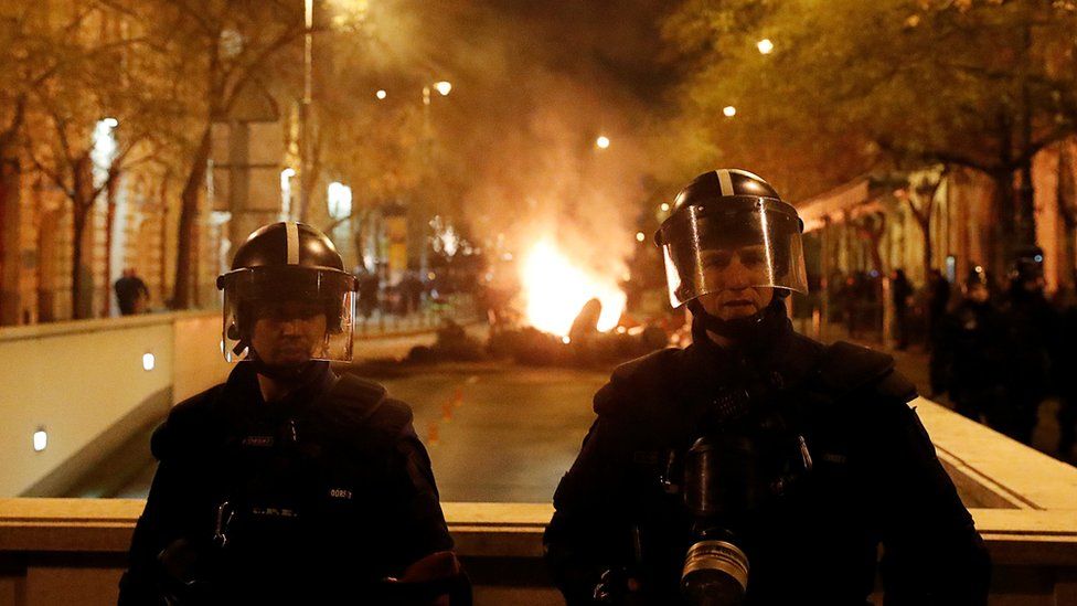 Two police in riot gear stand guard, while a fire burning in the street can be seen behind them