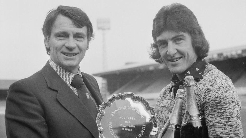 A black and white photo of Sir Bobby handing Brian Talbot a player of the month trophy in December 1976