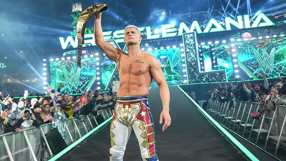 Cody Rhodes holds the winner's belt aloft after his victory. Cody is a 38-year-old white man with bleached blonde hair. He wears red, white and blue trousers and is shirtless, exposing his muscles. He's pictured surrounded by the WrestleMania audience in front of the event staging, which is lit green.