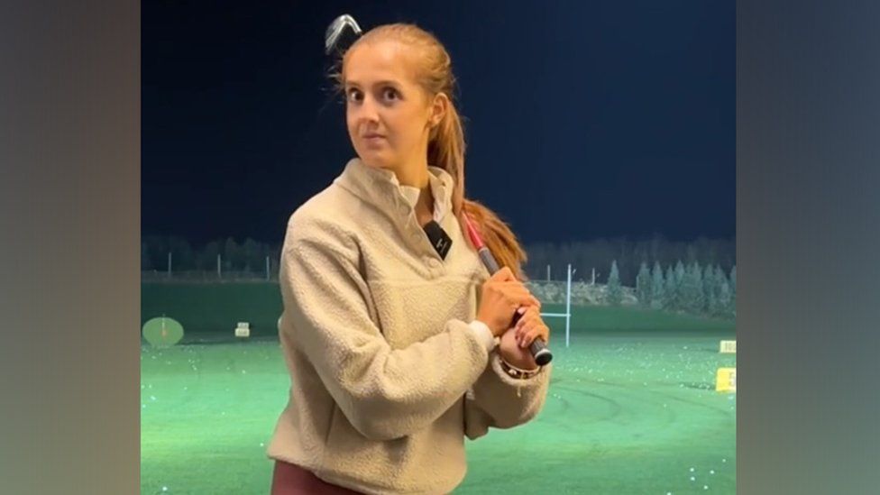 A young woman in a cream-coloured fleece and lilac leggings stands in front of a grass-covered driving range with a golf club resting on her shoulder. Her eyebrows are raised and she looks slightly shocked with a hint of amusement, like she can't quite believe what she just heard.