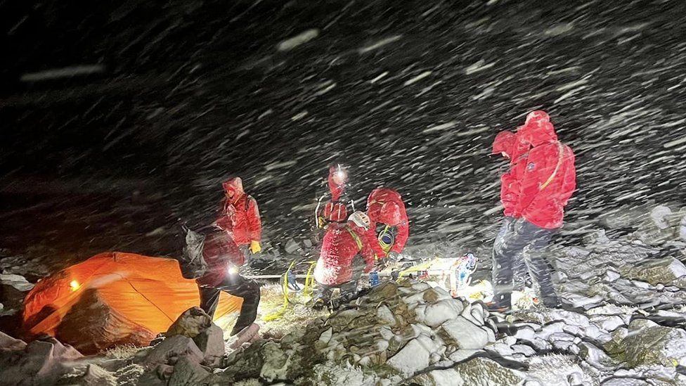 People in red jackets lid by headtorches with snow blowing around them attend to a casualty in a pop up orange tent