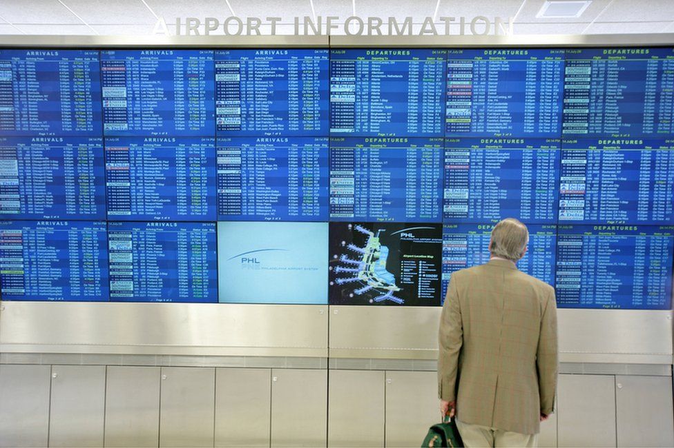 A person stands in front of a collection of airport information screens displaying arrivals and departures
