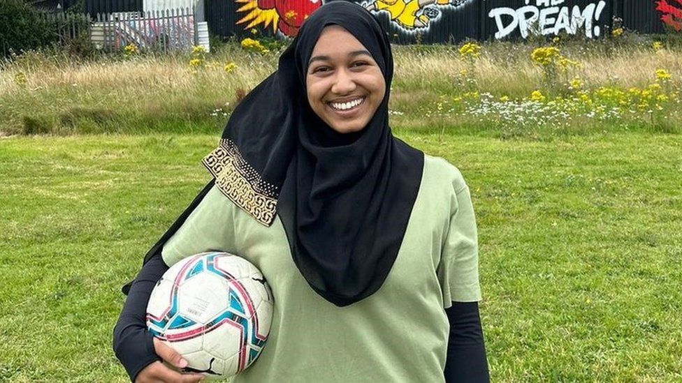 Yasmin Rahman standing in a grassy field holding a white football under her right arm. She is wearing a black hijab (headscarf) on her head and is smiling. She is wearing an olive green short-sleeved T-shirt and has a black long-sleeved shirt on underneath that. Behind her is a building with a mural on it that says "fulfilling the dream".